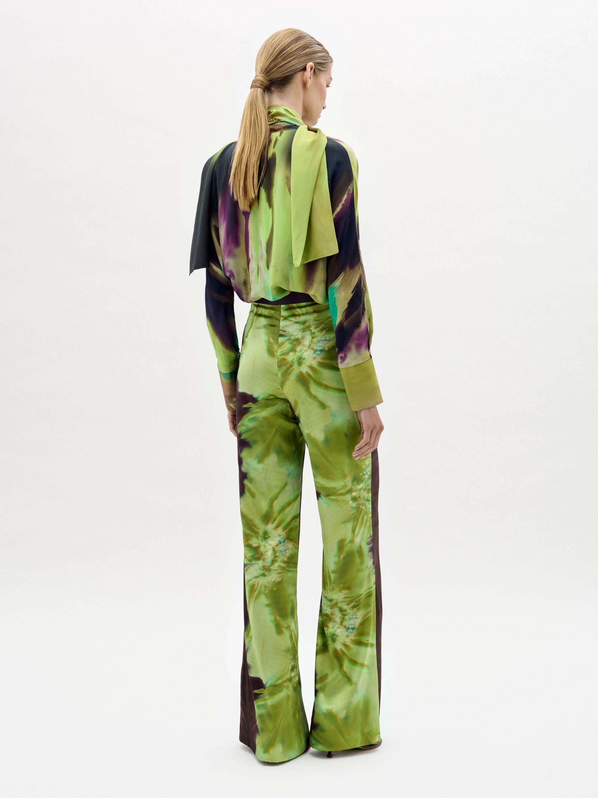A woman stands against a plain background wearing the Andie Pant, featuring a green and brown abstract print, paired with a matching blouse, wide belt, and scarf.