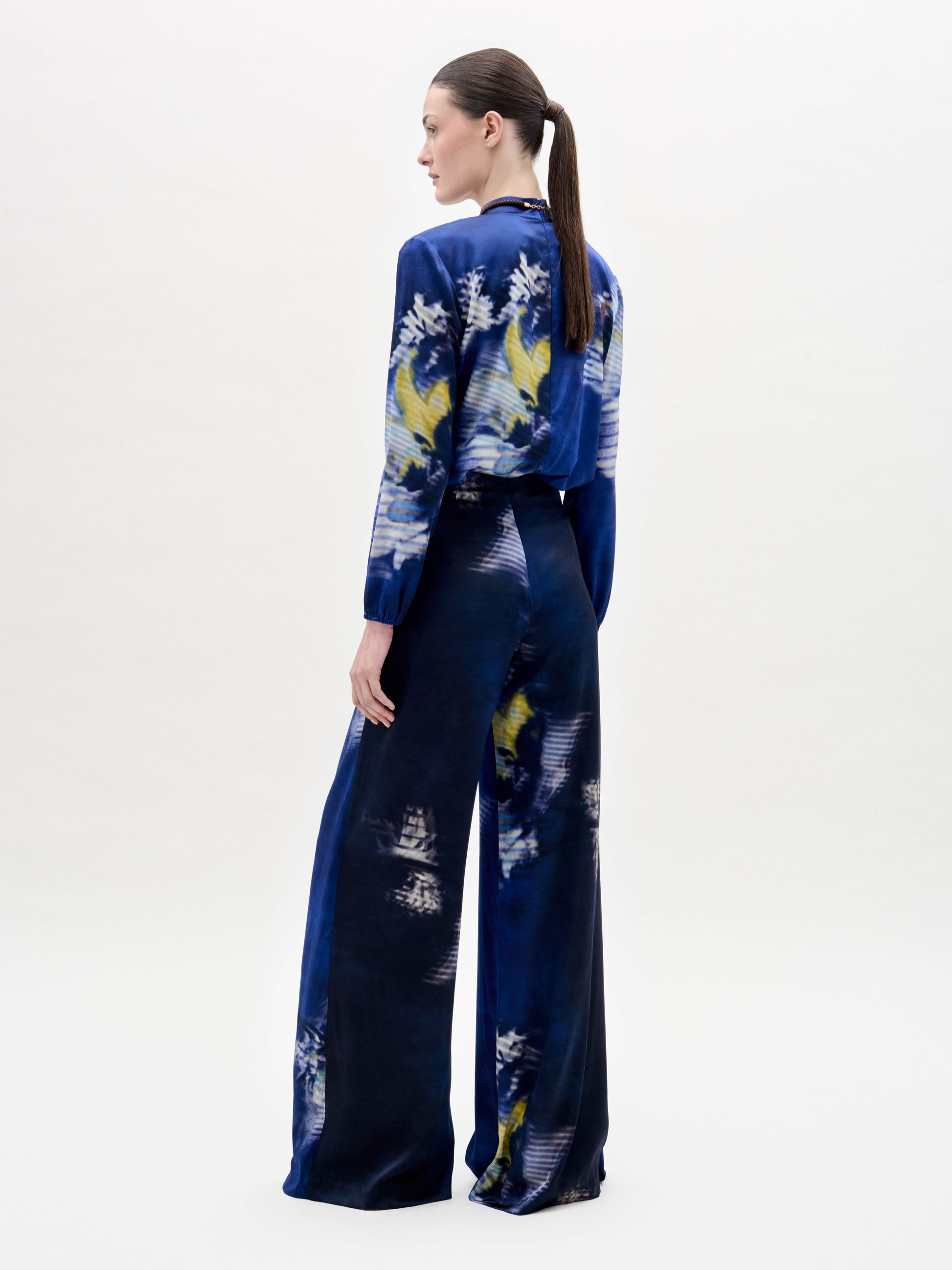 A person stands against a white background wearing the Canturipe Pant, paired with a blue patterned satin blouse and a choker-style scarf.