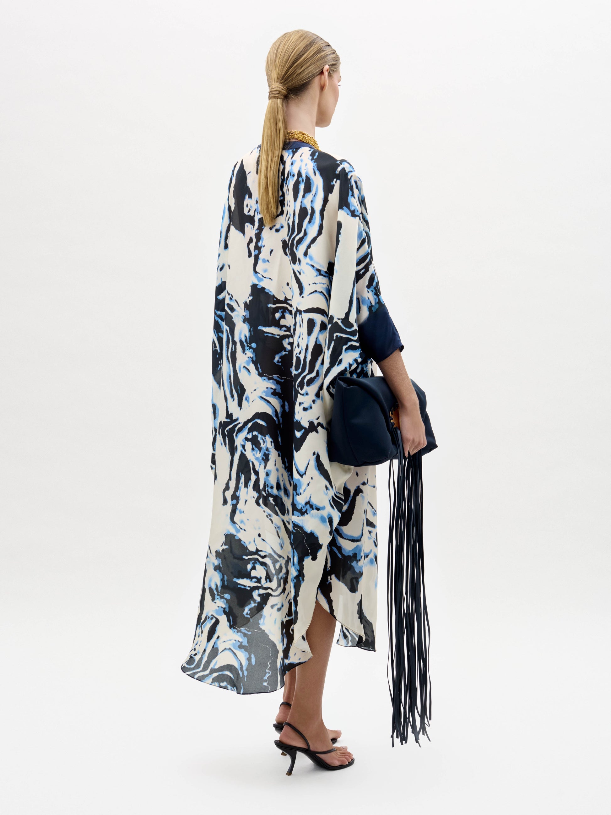 A woman stands against a plain background wearing the Elea Tunic with an abstract pattern, black sandals, gold jewelry, and holding a navy fringe bag.