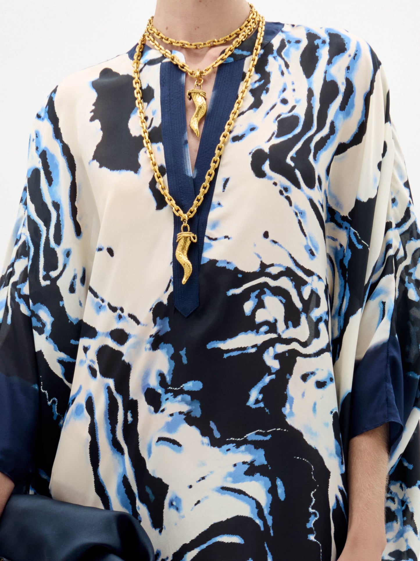 A woman stands against a plain background wearing the Elea Tunic with an abstract pattern, black sandals, gold jewelry, and holding a navy fringe bag.