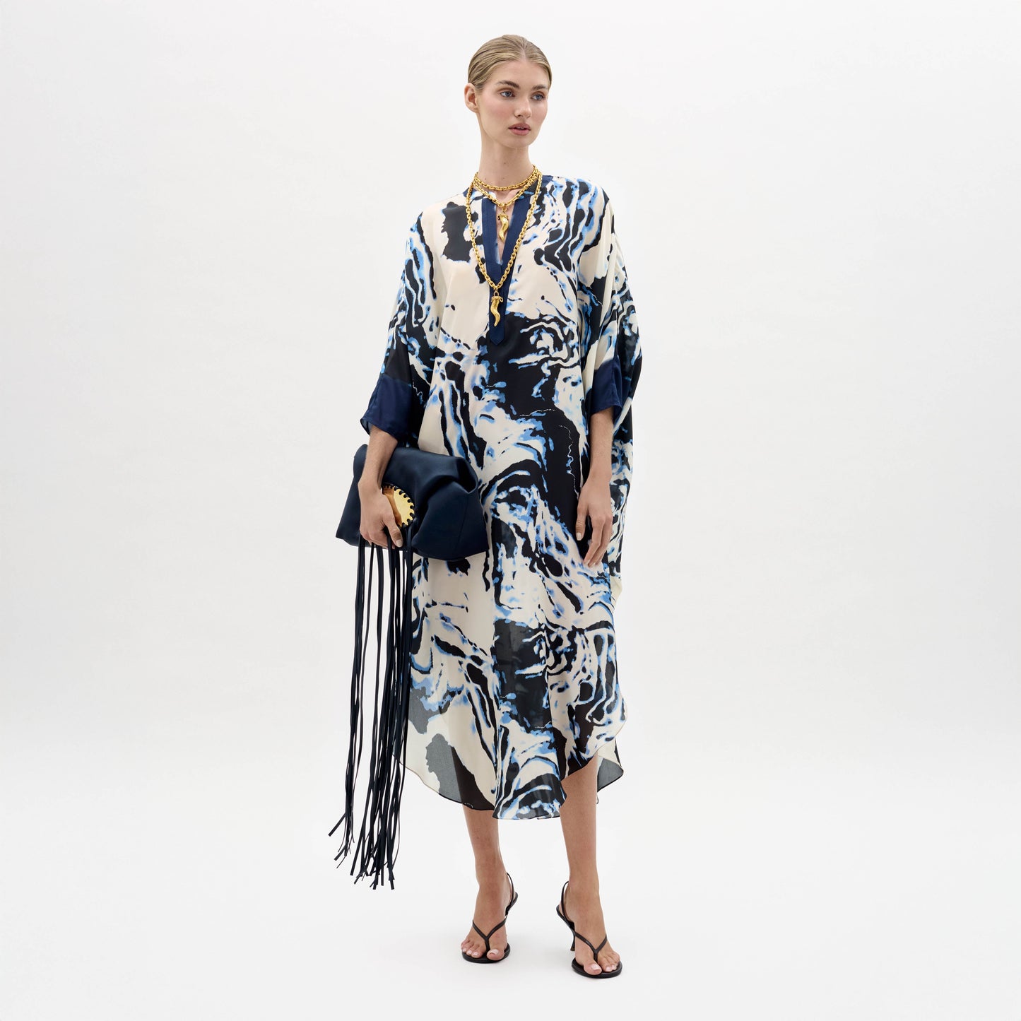 A woman stands against a plain background wearing the Elea Tunic with an abstract pattern, black sandals, gold jewelry, and holding a navy fringe bag.