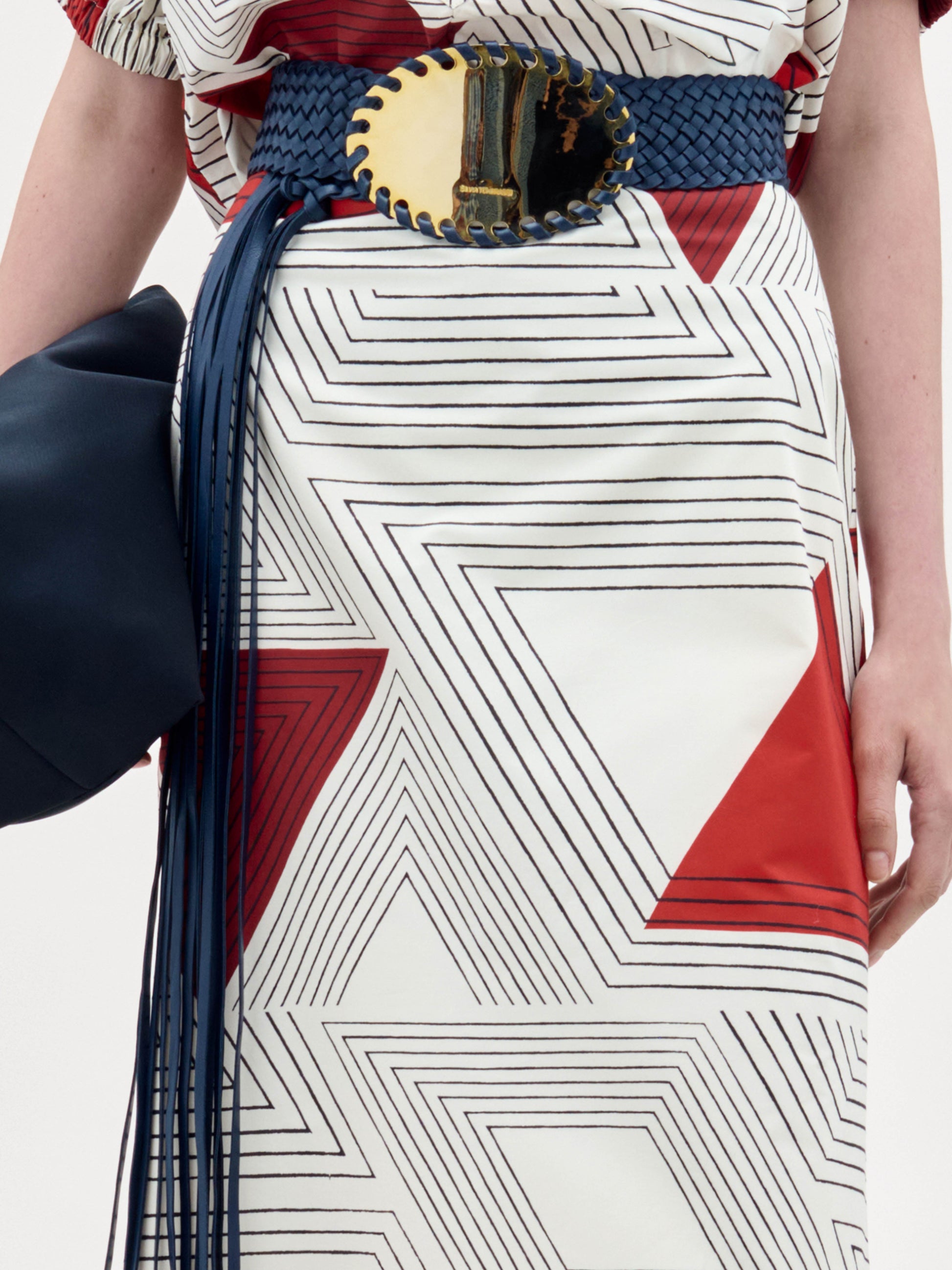 A woman stands against a plain background wearing the Atira Skirt paired with black sandals, a wide belt with a gold buckle, and holding a black bag at her side.