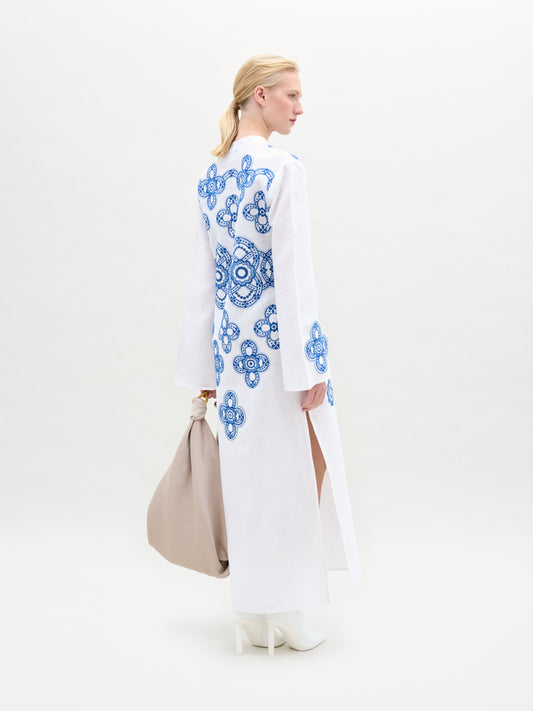 A woman stands against a white background wearing the Adalys Tunic, an Italian-fabric maxi with blue circular patterns and flared bell sleeves, styled with white boots and a large beige bag.