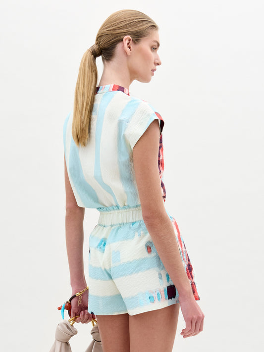 Wearing the Adele Blouse, a woman pairs its light blue and multicolored patterned collarless design with matching shorts made from Italian fabric, holding a small bag as she stands against a plain light background.
