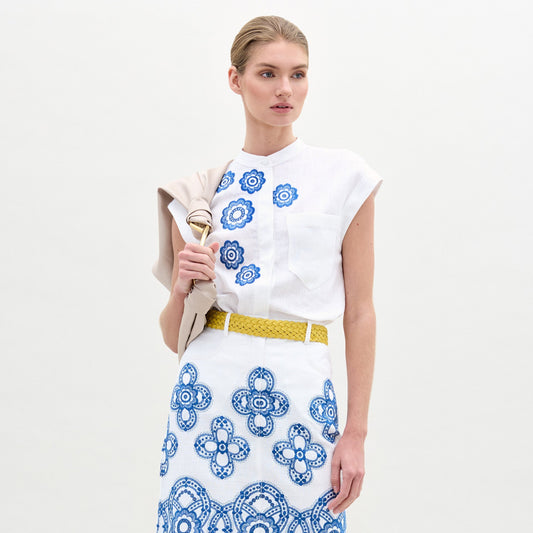 A woman wearing the Adele Blouse—a white, collarless top with blue floral motifs—and a matching skirt with a yellow belt stands against a plain background, a beige jacket draped over her shoulder.