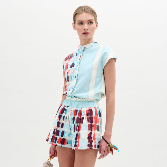 Wearing the Adele Blouse, a woman pairs its light blue and multicolored patterned collarless design with matching shorts made from Italian fabric, holding a small bag as she stands against a plain light background.