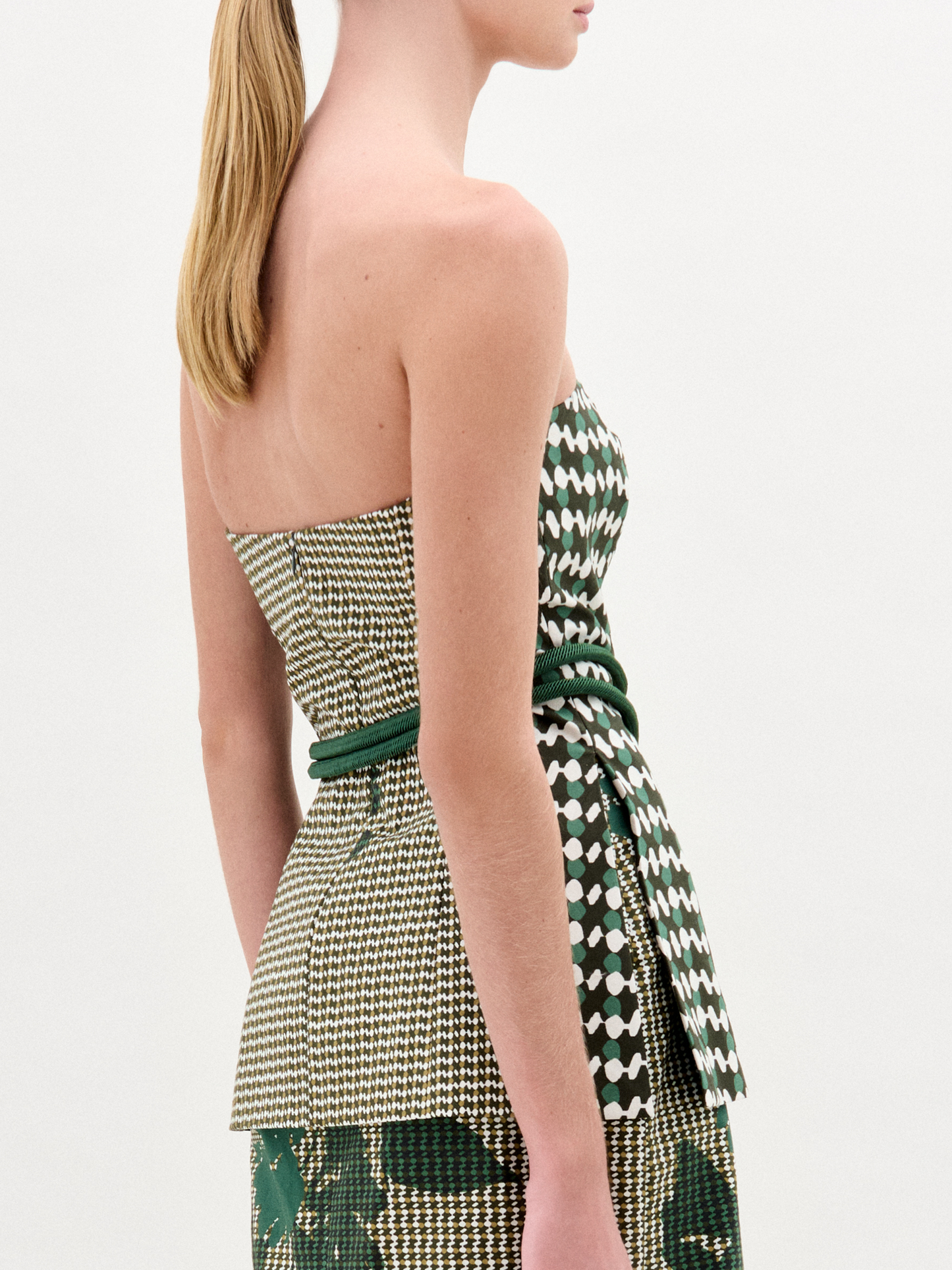 A woman wears the Astor Top, a strapless bustier in green, beige, and houndstooth Italian fabric with a matching skirt and a green belt with gold clasp, standing against a plain white background.