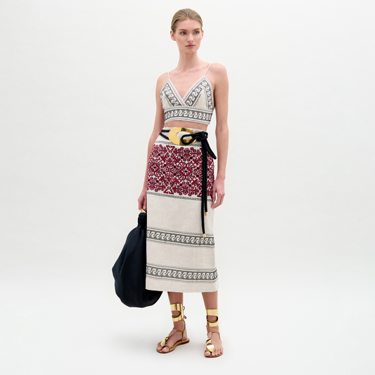 A woman stands against a plain background wearing the Atira Skirt, styled with a black belt, gold sandals, and carrying a large black bag.