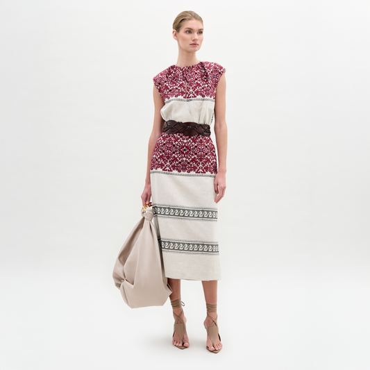 A woman stands against a white background wearing the Badra Dress, styled with a woven leather belt, beige strappy sandals, and holding a large beige bag.