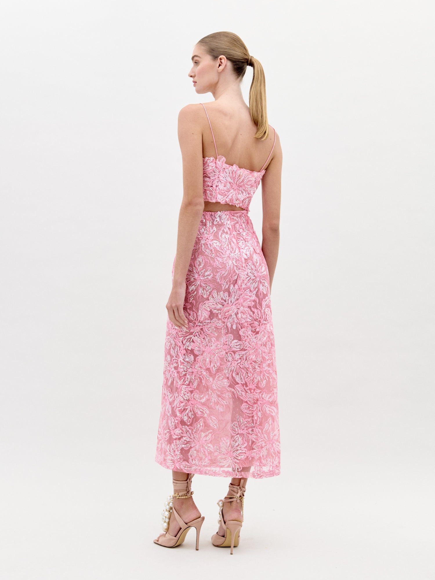 A woman stands wearing the Alida Dress Set Pink, a chic two-piece floral lace outfit featuring a crop top and a mid-length skirt, paired with high-heeled shoes adorned with decorative elements. Perfect for an elegant event, this set is guaranteed to impress; make sure to order by the ship-by date of February 1st, 2025.