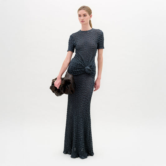 A woman poses against a plain background wearing the Ciji Skirt—crafted from premium Italian fabric, this dark, textured skirt features a side knot detail. She holds a dark clutch bag in her left hand.