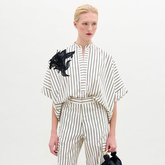 A person with blond hair in a low ponytail wears the Daia Blouse, a loose, white 100% cotton top with black vertical stripes and short sleeves, standing against a plain light background.