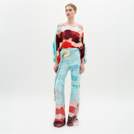 A woman stands against a plain light background wearing the Dalal Pant—high-waisted, wide-leg pants in luxurious Italian fabric with an abstract pattern—styled with a colorful off-the-shoulder top and finished with a yellow belt accent.