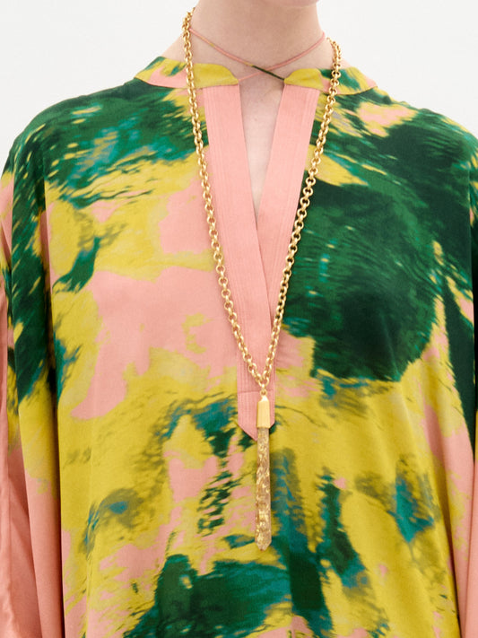 A woman stands against a white background wearing the Elea Tunic, an oversized 100% silk maxi dress with pink, yellow, and green patterns, paired with a beige bag and strappy heels.
