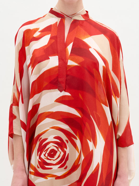 A woman stands against a white background wearing the Elea Tunic, a loose, knee-length dress made from Italian fabric with a red and cream spiral pattern. She holds a beige clutch and pairs the look with gold high-heeled sandals.