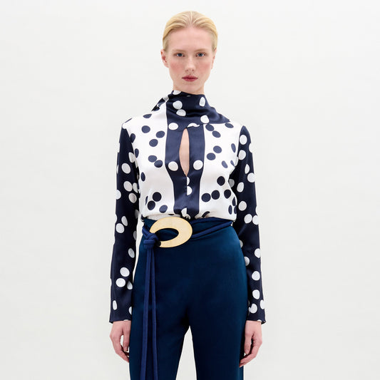 A woman stands against a plain background, wearing the Gaby Blouse—a navy and white polka dot silk top with a keyhole neckline—paired with high-waisted navy pants featuring a large oval belt buckle.