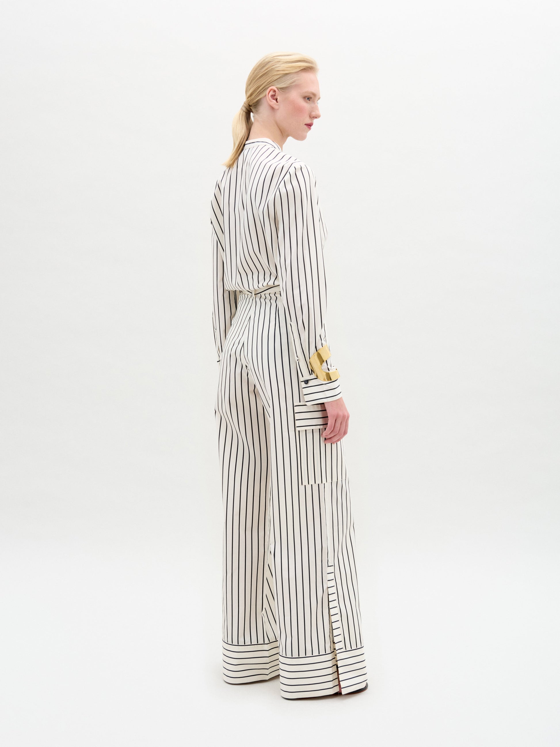 A person stands against a white background wearing a white long-sleeved blouse with vertical black stripes and lace detail, paired with the Falak Pant—relaxed fit, wide-leg trousers crafted from premium Italian fabric.