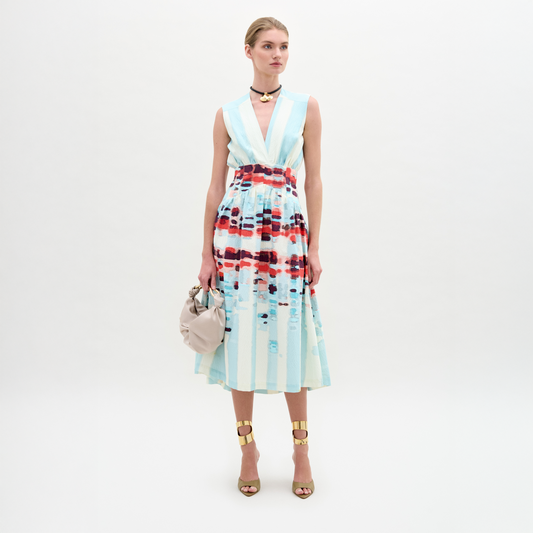 A woman poses against a plain backdrop in the Jocelyn Dress, a sleeveless V-neck midi made from fine Italian fabric with blue stripes and red patterns. She accents the look with gold sandals, a choker, and carries a beige handbag.