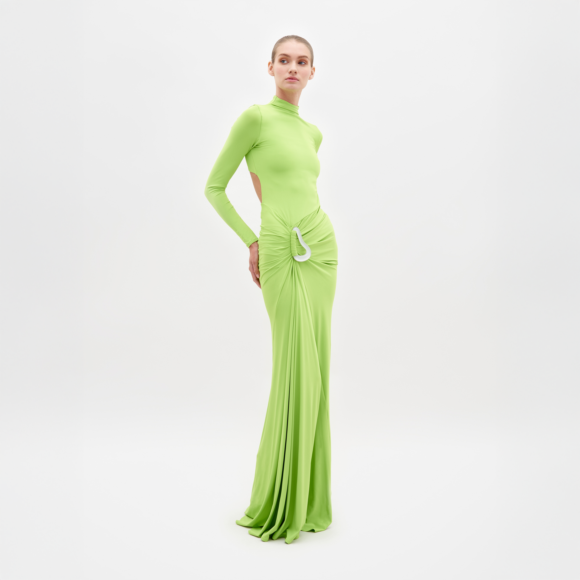A person is wearing the Lena Dress Lime, a long, lime green dress with long sleeves and a high neckline. The dress, which is available for shipping by February 15th, features cut-out details at the sides and a gathered, draped front. White background.