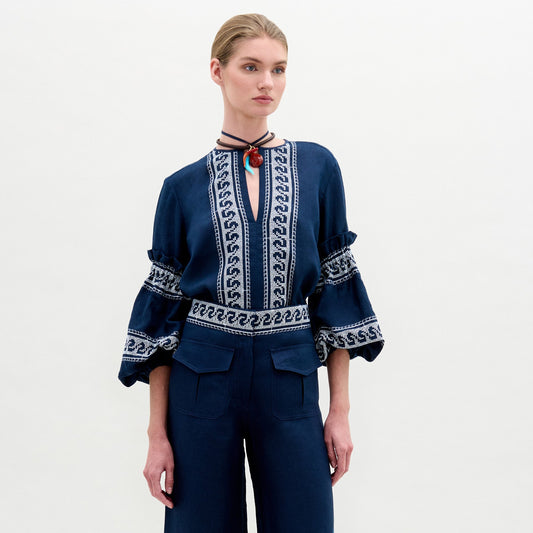 A woman stands against a plain white background wearing the Lucaya Blouse—an embroidered navy blue linen top with voluminous sleeves—paired with matching navy trousers.