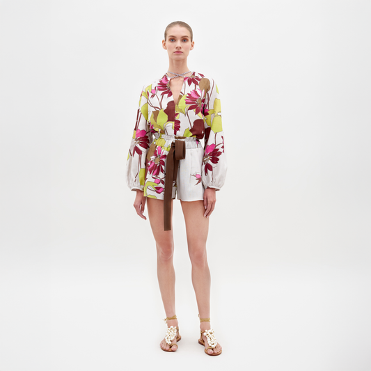 A person stands against a white background wearing the Giorgio Short Botanical Multifloral blouse, white shorts with a brown belt, and sandals. Pre-order this stylish ensemble now to ensure it arrives in time for February 15th, 2025.