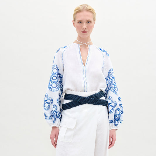 A woman stands against a plain background wearing the Molveno Blouse, an embroidered linen top with blue detailing and a V neckline, paired with a white skirt featuring a pocket and finished with a dark braided belt at her waist.