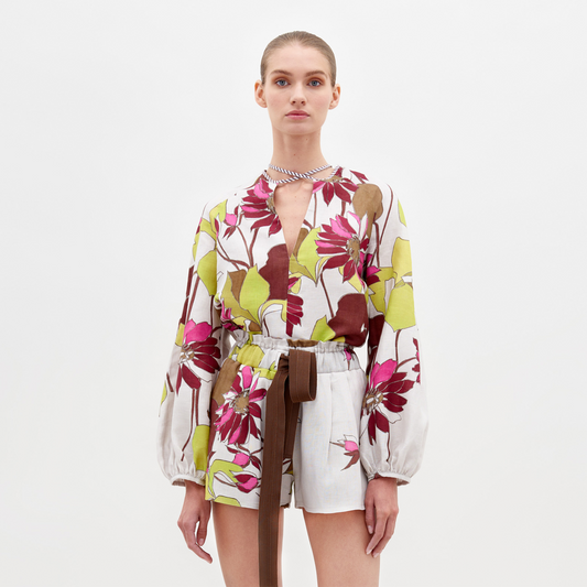 A person stands against a plain background, wearing the Molveno Blouse in Botanical Multifloral, featuring a floral-patterned long-sleeve top with a deep V-neckline and matching high-waisted shorts, cinched at the waist with a brown belt. Pre-order now to ensure it arrives by the February 15th, 2025 ship-by date.