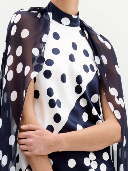 In a studio, a person models the Sabina Dress—a long black silk maxi with white polka dots and a matching cape overlay—paired with black and gold sandals.