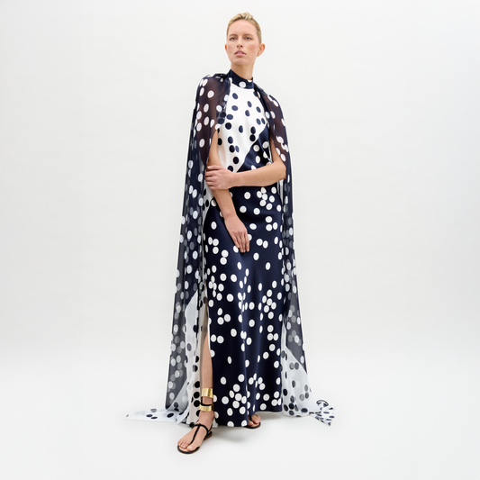 In a studio, a person models the Sabina Dress—a long black silk maxi with white polka dots and a matching cape overlay—paired with black and gold sandals.