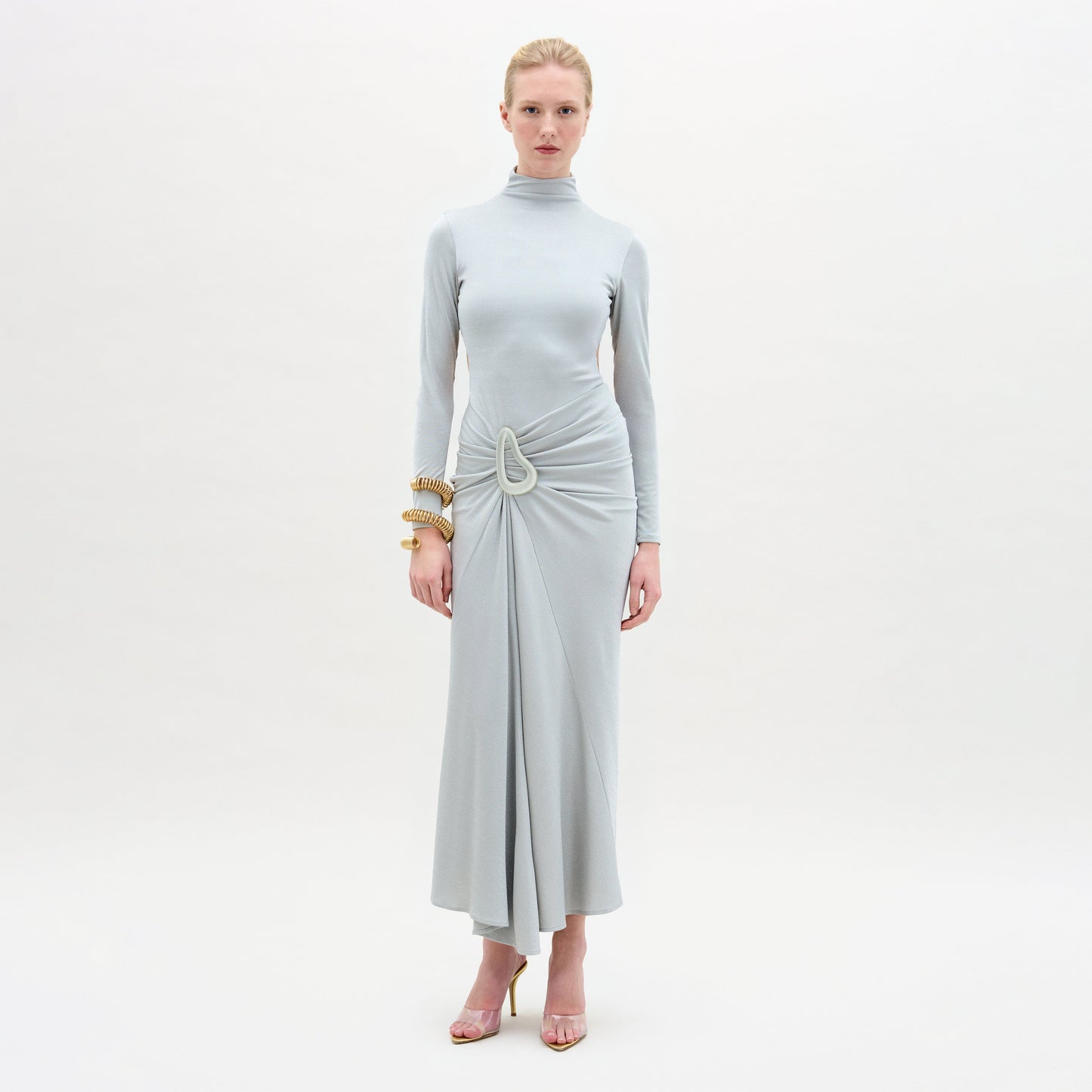 A woman poses against a plain white background, wearing the Venus Dress—a long-sleeved, light gray midi dress with a high neckline and draped skirt in fine Italian fabric—styled with gold bracelets and strappy high-heeled sandals.