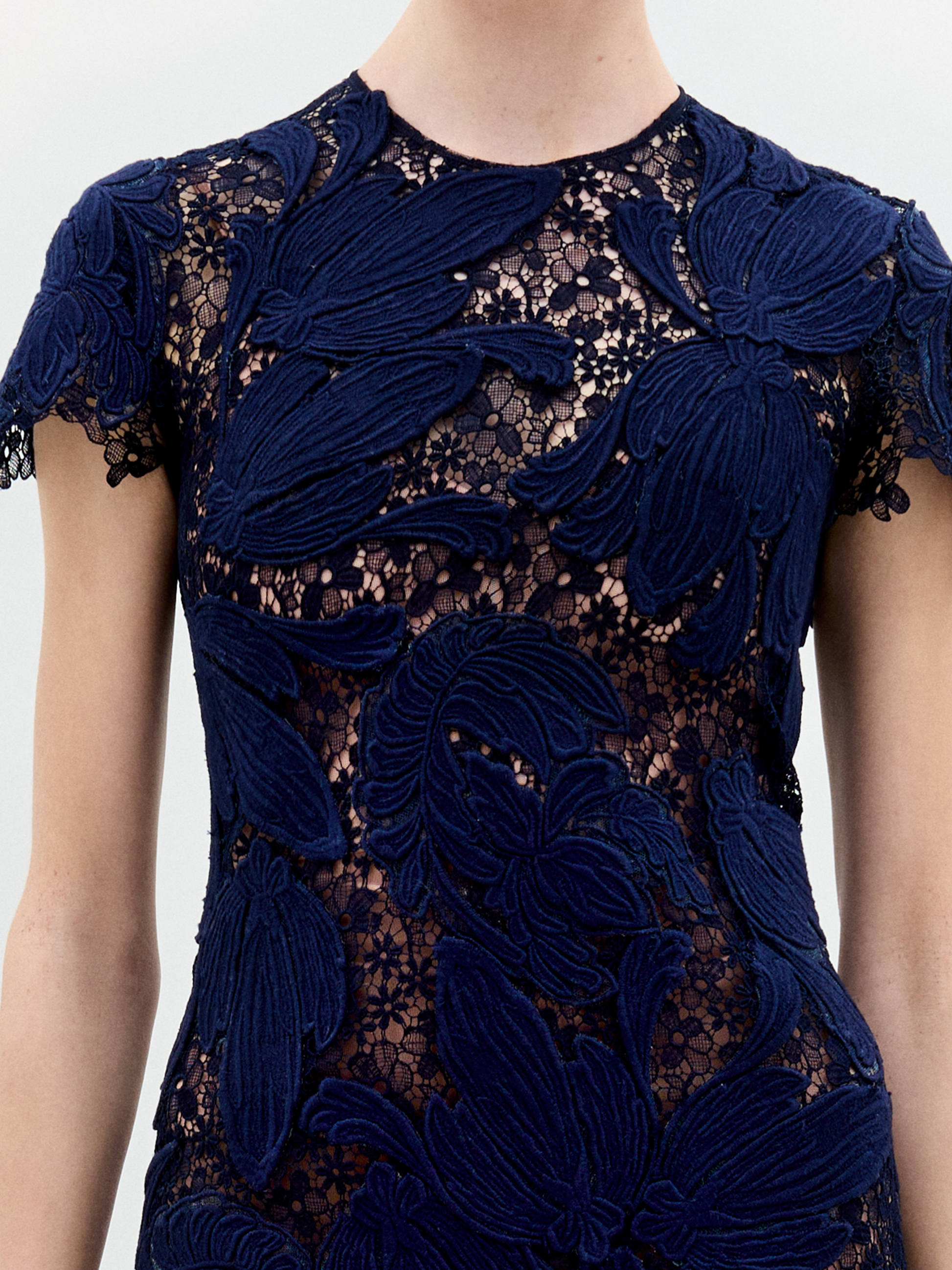 A person wears the Veronica Dress, a navy blue short-sleeved top made of sheer lace with large floral appliqué patterns that subtly reveal some skin. The photo focuses on the upper torso, with the face out of frame.