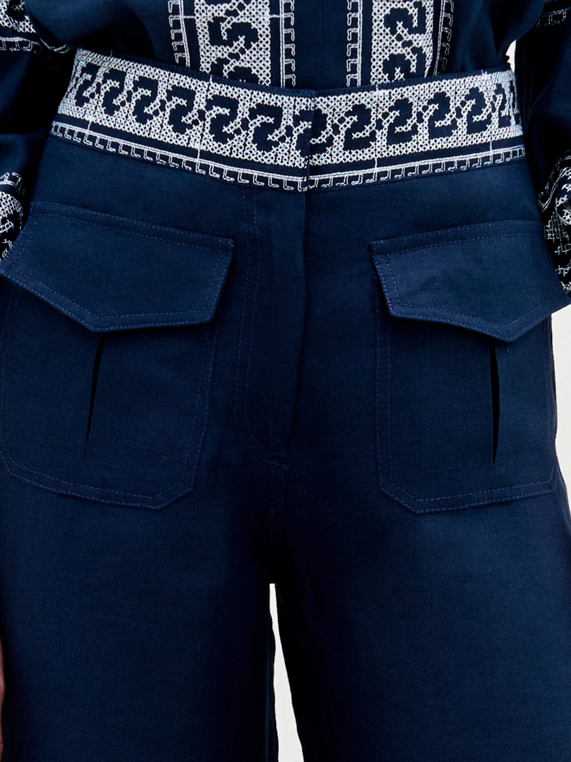 A woman stands against a plain background, wearing a navy blue embroidered blouse with ruffled sleeves and the Zina Pant—matching embroidered linen wide-leg pants—paired with gold sandals.